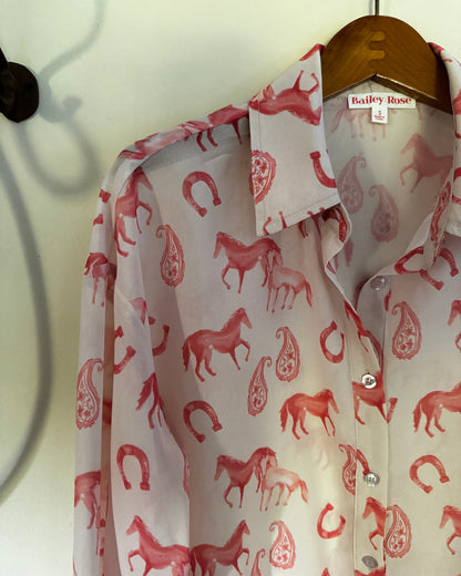 A Bailey Rose button-down shirt with a red and pink horse and paisley pattern, hanging on a wooden hanger against a light wall.