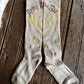A pair of ribbed beige crew socks laid flat on a dark wooden plank surface. The socks feature yellow graphic print and the phrase 'Enjoy The Dirt' in red script near the cuff, with two small yellow flower embroidery details on the sides.