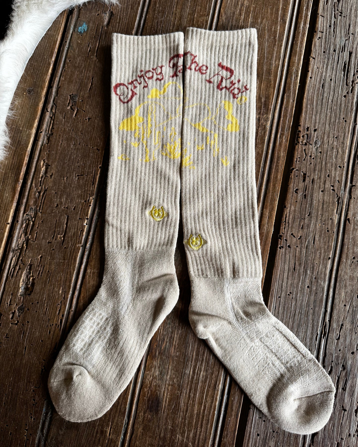 A pair of ribbed beige crew socks laid flat on a dark wooden plank surface. The socks feature yellow graphic print and the phrase 'Enjoy The Dirt' in red script near the cuff, with two small yellow flower embroidery details on the sides.