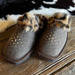 A pair of women's slip-on boots in dark brown felt with leopard-print faux fur trim and gold star-shaped studs, displayed on a rustic wooden floor over a black-and-white cowhide rug.