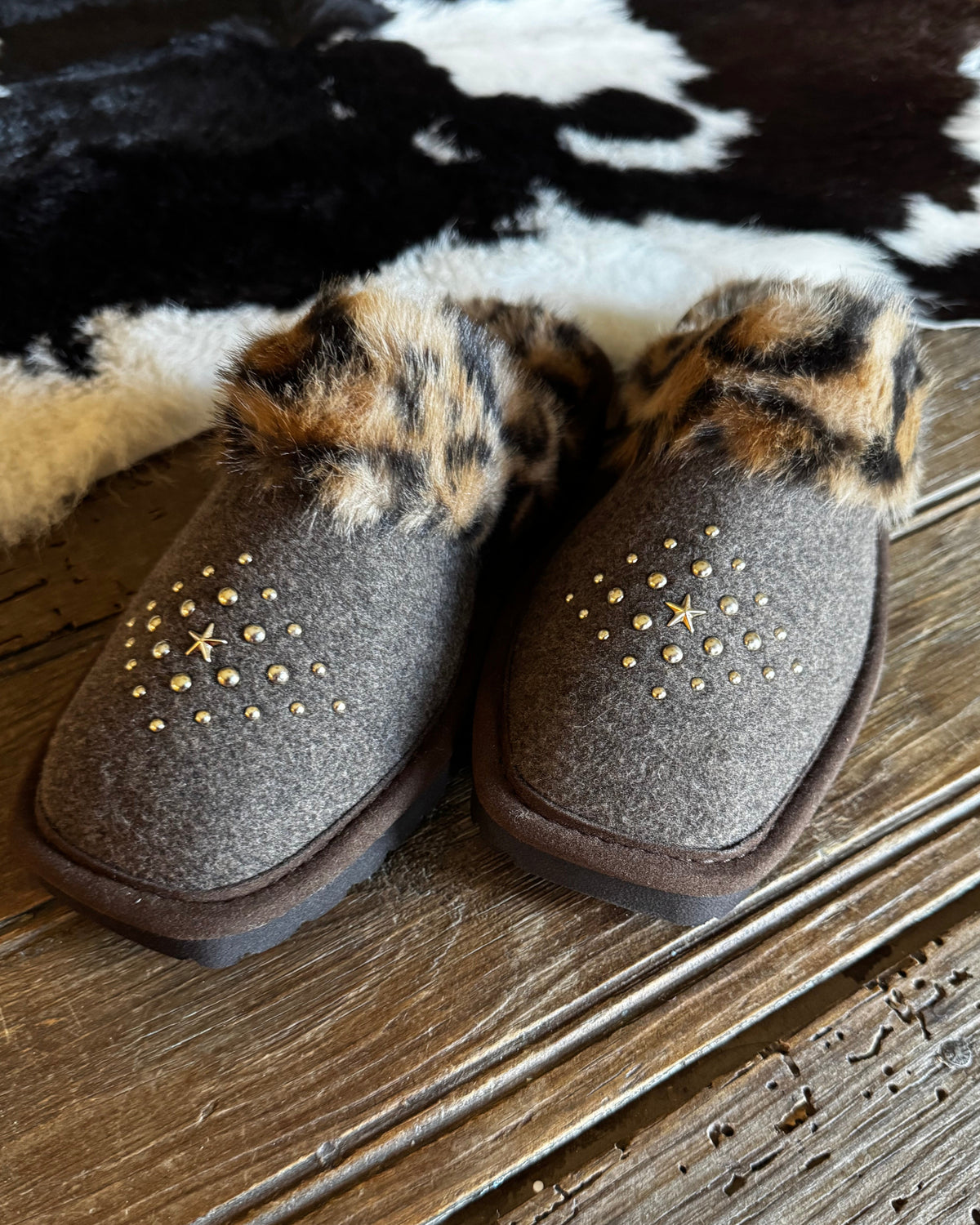 A pair of women's slip-on boots in dark brown felt with leopard-print faux fur trim and gold star-shaped studs, displayed on a rustic wooden floor over a black-and-white cowhide rug.
