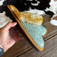 A hand holds a pair of western-style slippers with a turquoise tooled leather upper, tan suede trim, and a fluffy cream shearling lining, set against a rustic cowhide rug and wooden floor.
