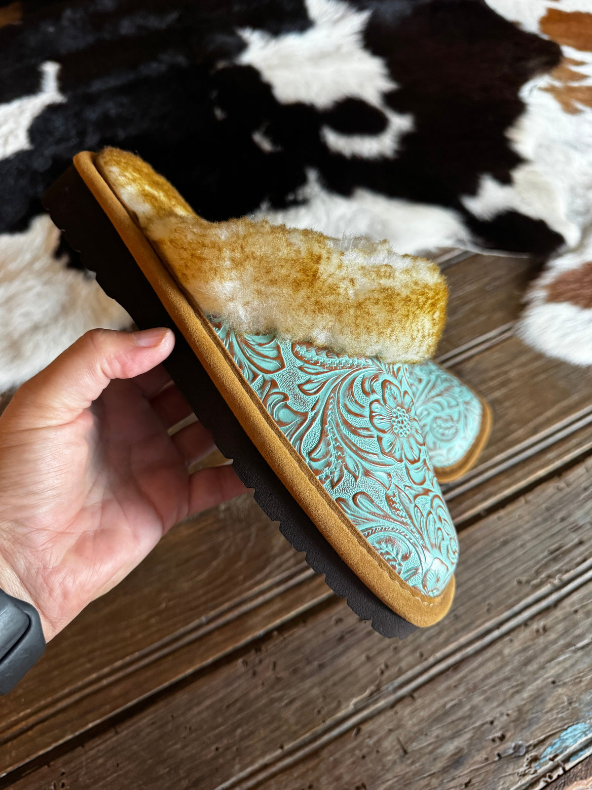 A hand holds a pair of western-style slippers with a turquoise tooled leather upper, tan suede trim, and a fluffy cream shearling lining, set against a rustic cowhide rug and wooden floor.
