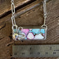 A silver-toned rectangular pendant necklace with a colorful marbled inlay of pink, blue, white, and brown, displayed on a rustic wooden surface next to a 2-inch ruler for scale.
