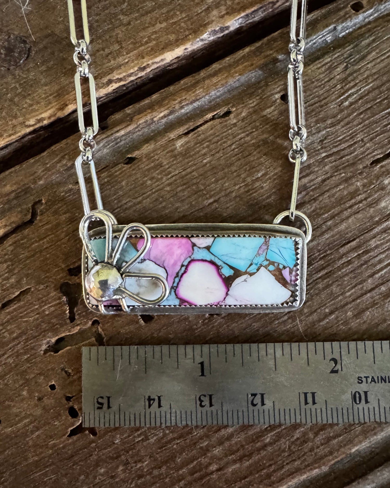 A silver-toned rectangular pendant necklace with a colorful marbled inlay of pink, blue, white, and brown, displayed on a rustic wooden surface next to a 2-inch ruler for scale.