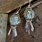 A pair of handmade silver earrings with circular textured stone settings and dangling silver leaf charms, shown next to a metal ruler on a dark wooden surface.