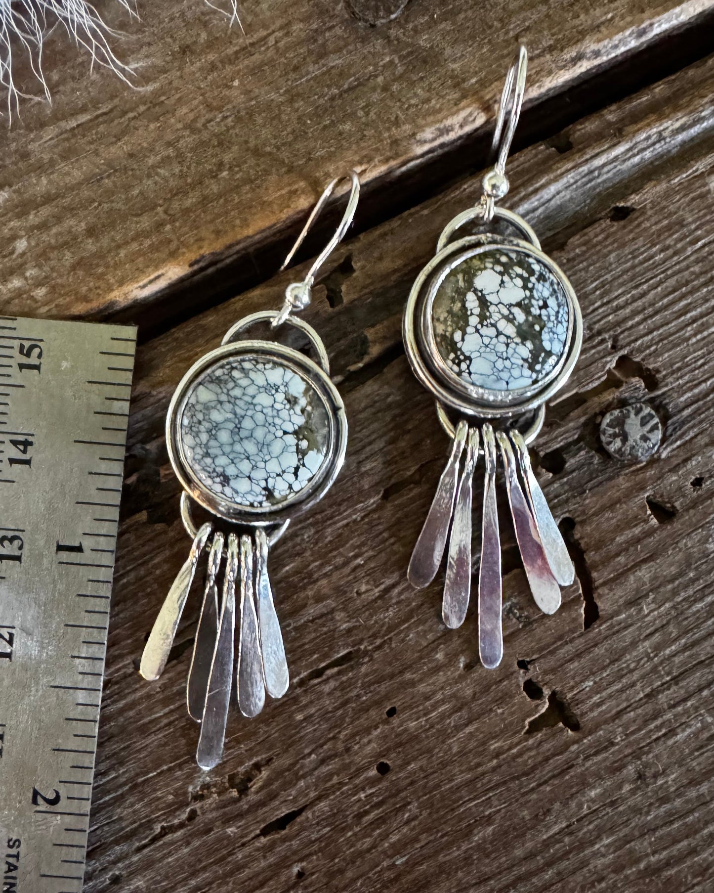 A pair of handmade silver earrings with circular textured stone settings and dangling silver leaf charms, shown next to a metal ruler on a dark wooden surface.