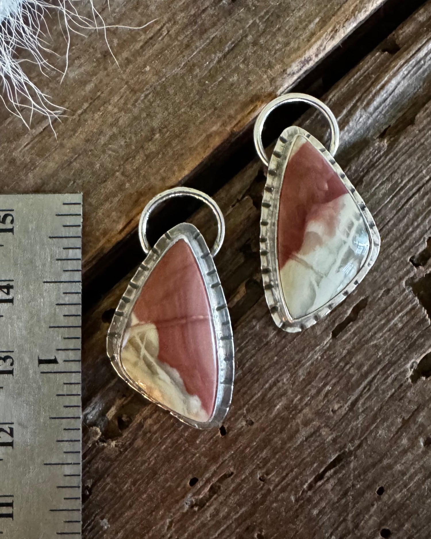 A pair of teardrop-shaped dangle earrings with pinkish-brown and cream marbled stones in silver bezel settings, displayed on a rustic wooden surface next to a metal ruler for scale.