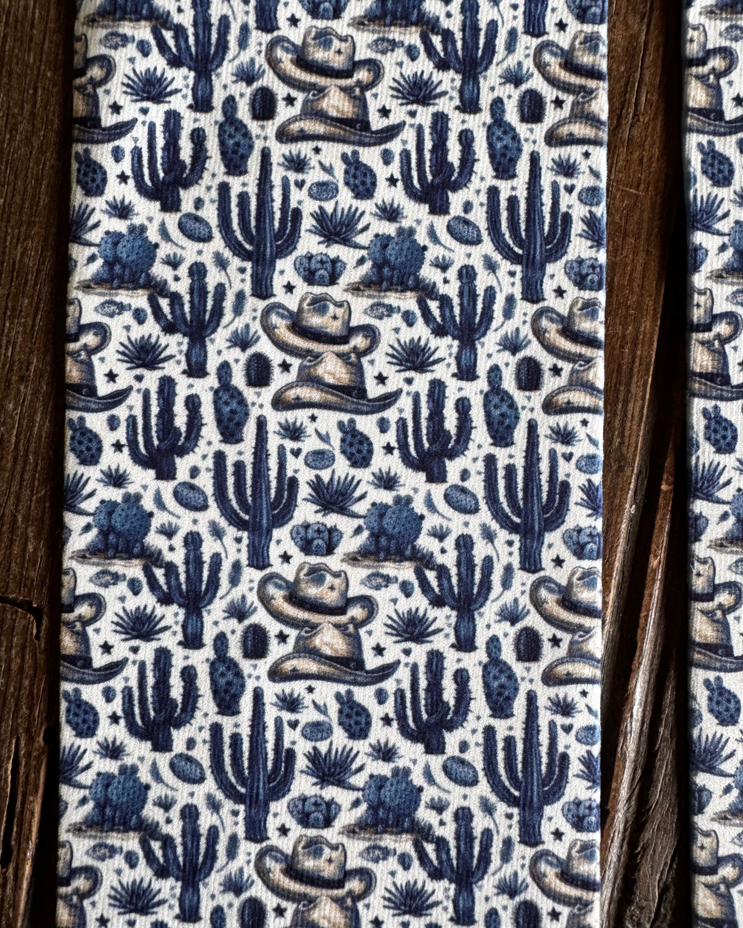 A close-up of a patterned fabric featuring a blue and beige cowboy and cactus motif on a white background, framed by dark brown wooden boards on the sides.