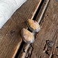 A pair of silver-toned teardrop-shaped earrings with textured, speckled yellow and brown stone inlays, displayed on a rustic dark wood surface next to a white faux fur trim.