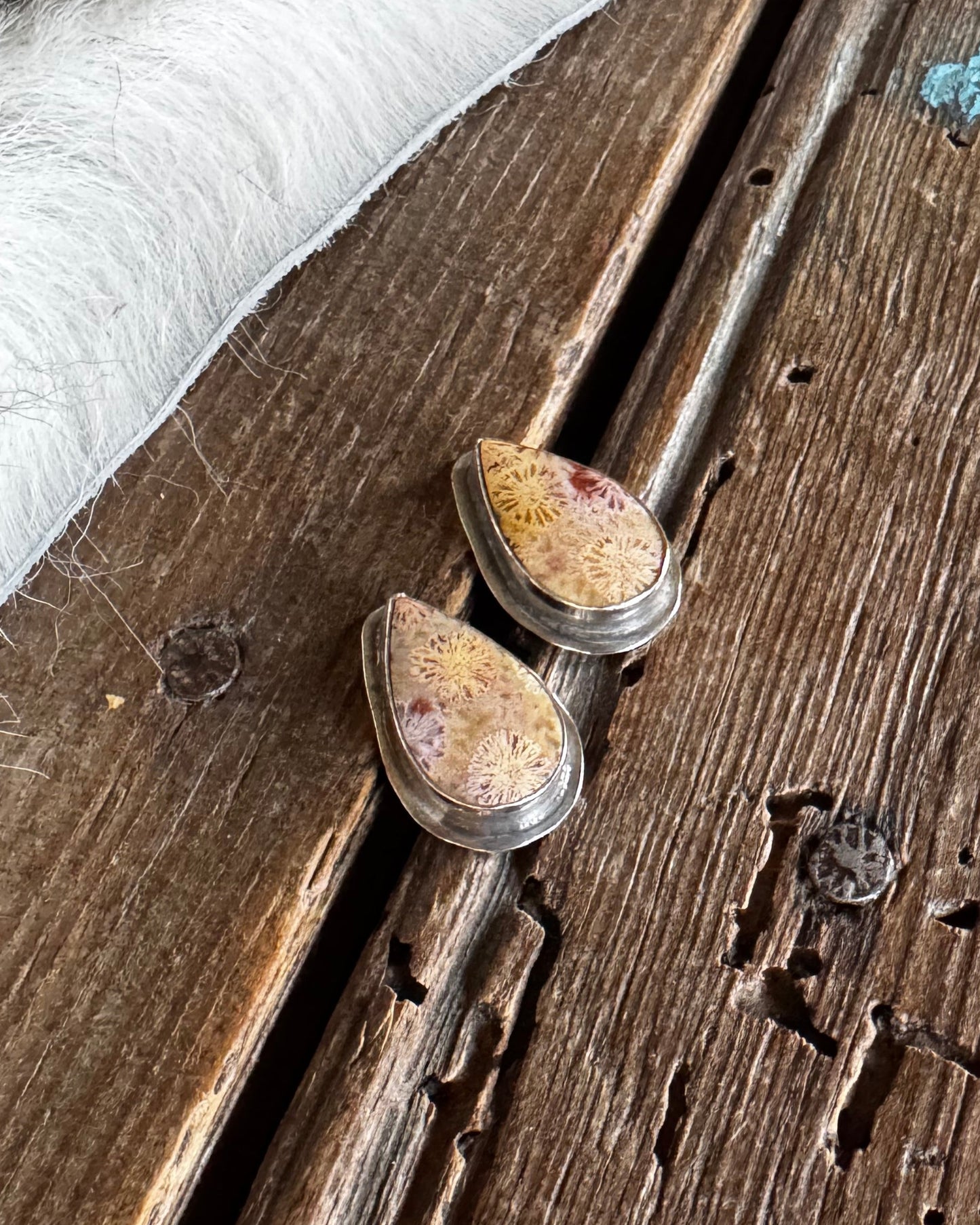 A pair of silver-toned teardrop-shaped earrings with textured, speckled yellow and brown stone inlays, displayed on a rustic dark wood surface next to a white faux fur trim.
