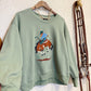 A sage green crewneck sweatshirt hanging on a wooden hanger against a textured white wall. The sweatshirt features a graphic print of a cowboy riding a brown horse.