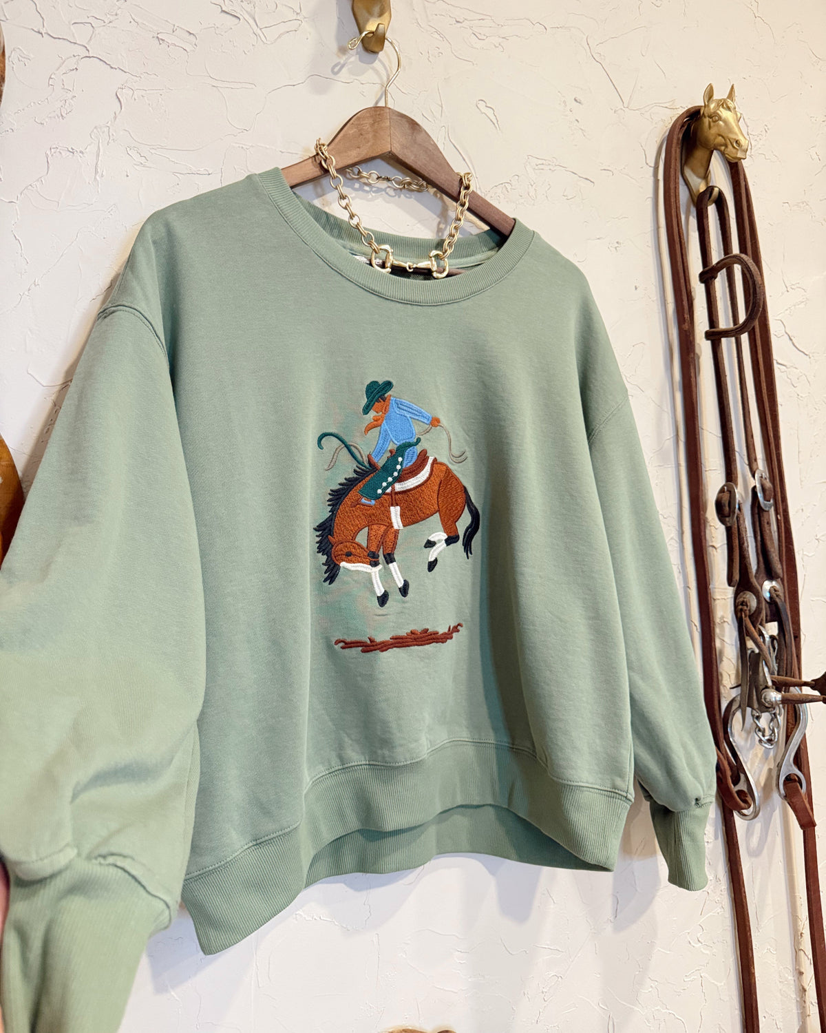 A sage green crewneck sweatshirt hanging on a wooden hanger against a textured white wall. The sweatshirt features a graphic print of a cowboy riding a brown horse.