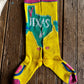 A pair of bright yellow ribbed crew socks laid flat on a rustic dark wood surface. The socks feature a large green map-shaped graphic with 'TEXAS' in white across the top, pink cuffs and toes, and blue and pink heel details.