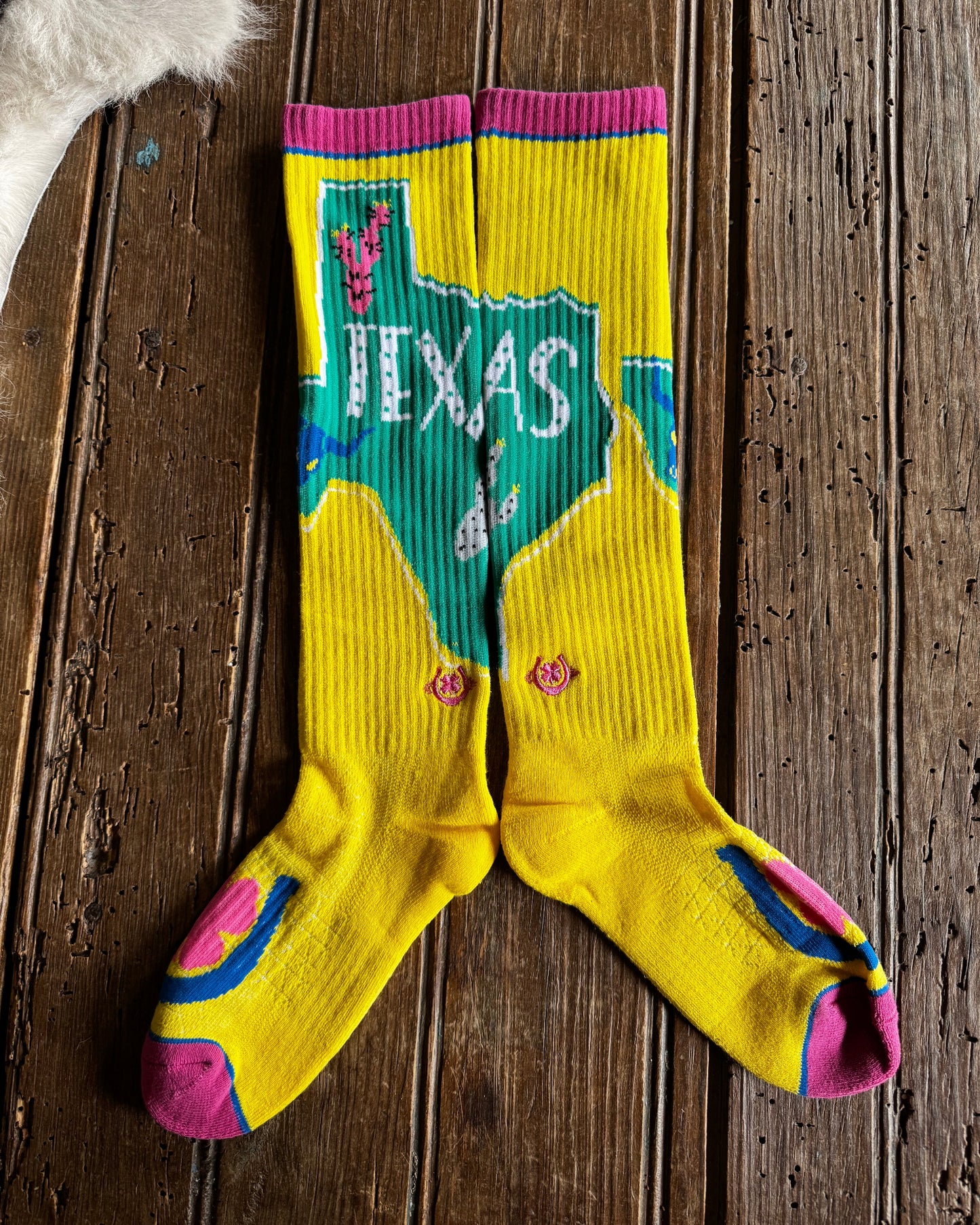 A pair of bright yellow ribbed crew socks laid flat on a rustic dark wood surface. The socks feature a large green map-shaped graphic with 'TEXAS' in white across the top, pink cuffs and toes, and blue and pink heel details.