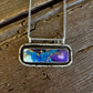 A silver-toned chain necklace with a rectangular bezel pendant featuring a marbled blue, purple, and white design, displayed on a rustic dark wood background.