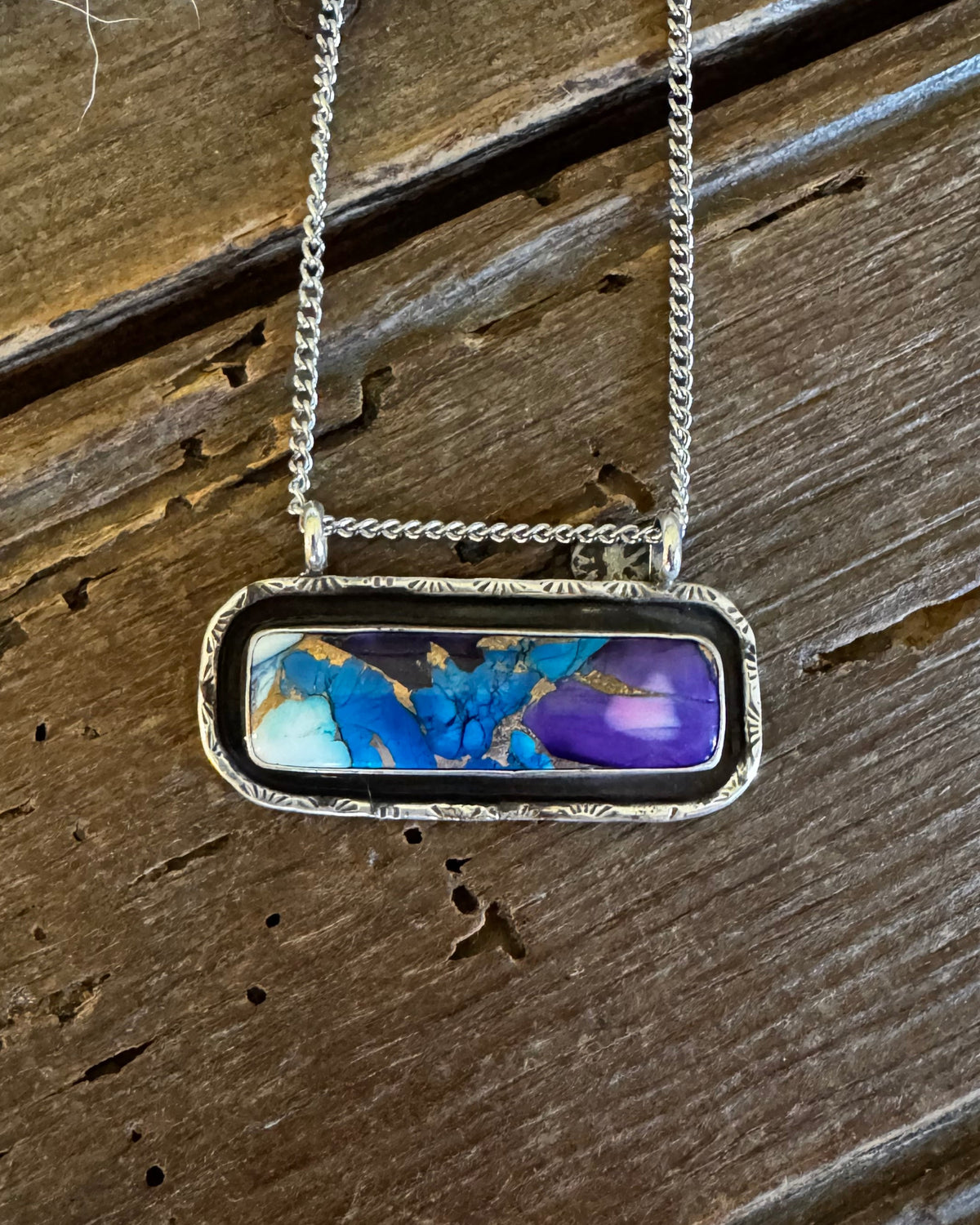 A silver-toned chain necklace with a rectangular bezel pendant featuring a marbled blue, purple, and white design, displayed on a rustic dark wood background.