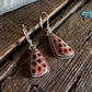 A pair of handmade dangle earrings with triangular coral-like pendants featuring a circular pattern, suspended from silver fishhook wires, displayed on a rustic dark wood surface with a white faux fur accent.