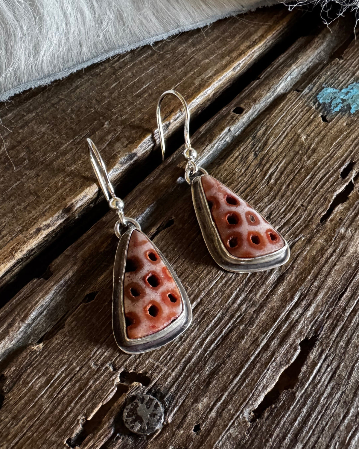 A pair of handmade dangle earrings with triangular coral-like pendants featuring a circular pattern, suspended from silver fishhook wires, displayed on a rustic dark wood surface with a white faux fur accent.