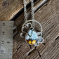 A silver-toned necklace with a circular filigree pendant and a teardrop-shaped stone featuring a blue-gray matrix with bright yellow and orange inclusions, displayed on a rustic wooden surface next to a metal ruler for scale.