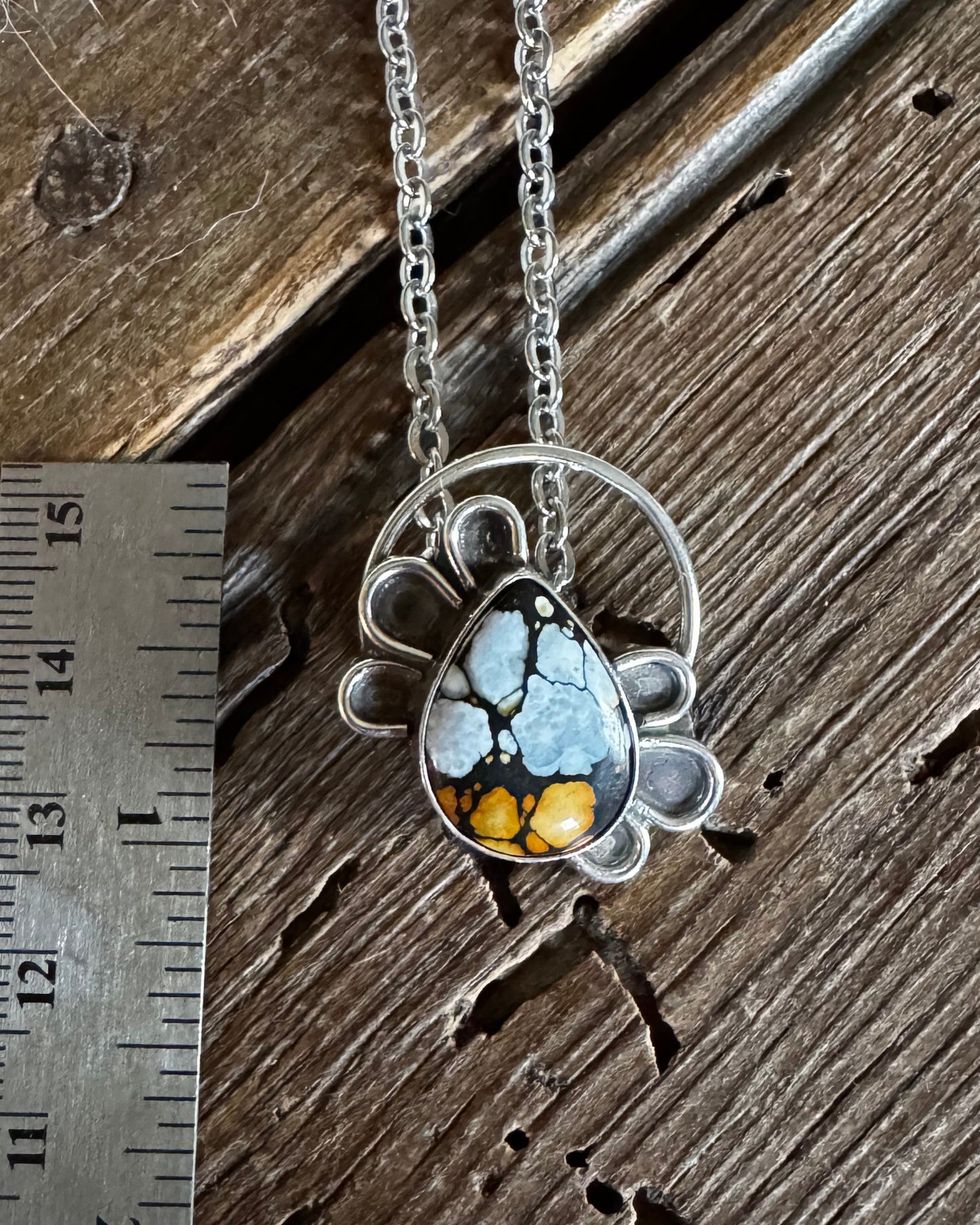 A silver-toned necklace with a circular filigree pendant and a teardrop-shaped stone featuring a blue-gray matrix with bright yellow and orange inclusions, displayed on a rustic wooden surface next to a metal ruler for scale.