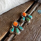 A pair of handmade silver dangle earrings with bright orange cabochon stones and turquoise teardrop accents, displayed on a rustic wooden surface next to a white faux fur trim.