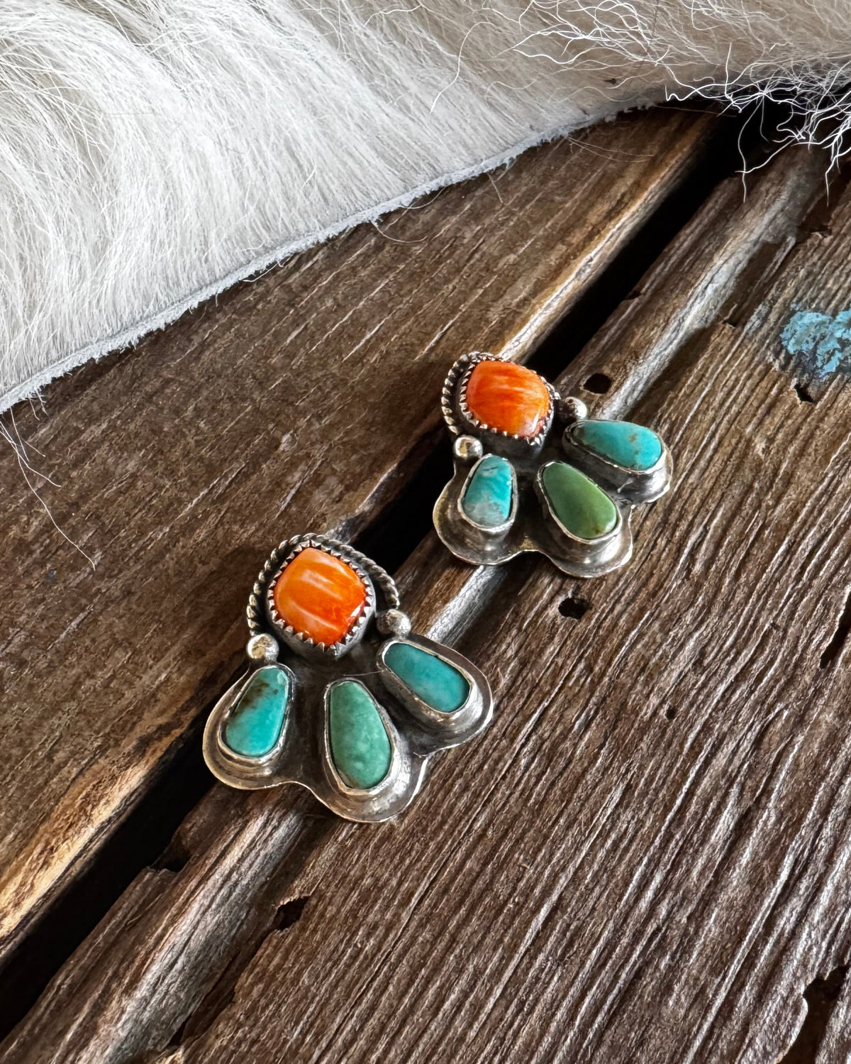 A pair of handmade silver dangle earrings with bright orange cabochon stones and turquoise teardrop accents, displayed on a rustic wooden surface next to a white faux fur trim.