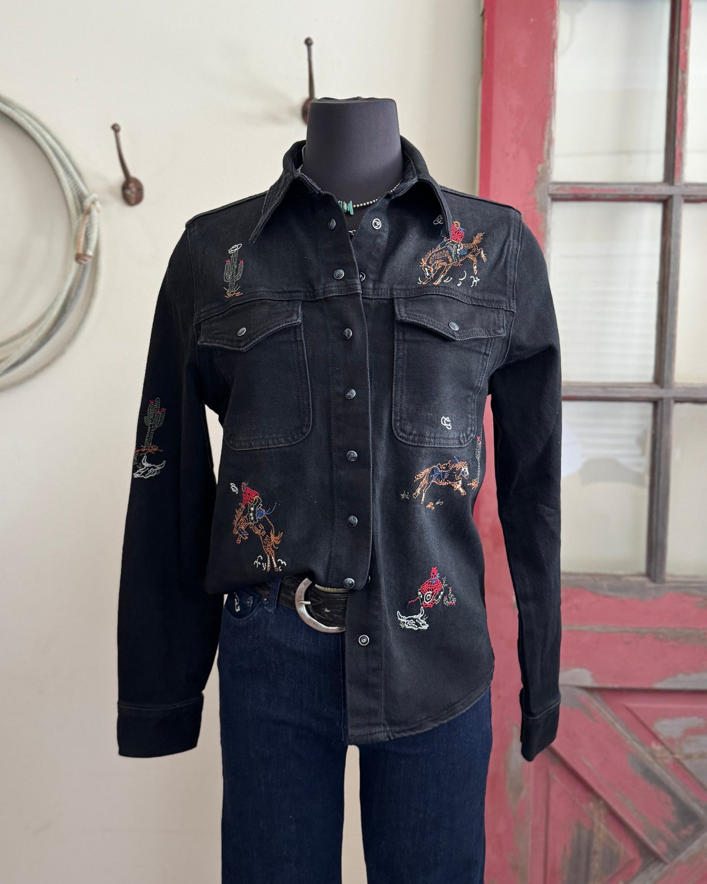 Black denim shirt with embroidery on a mannequin against a rustic background.