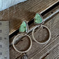 A pair of silver-toned dangle earrings with triangular light green gemstone tops and textured circular wire hoops, displayed on a rustic wooden surface next to a metal ruler for scale.