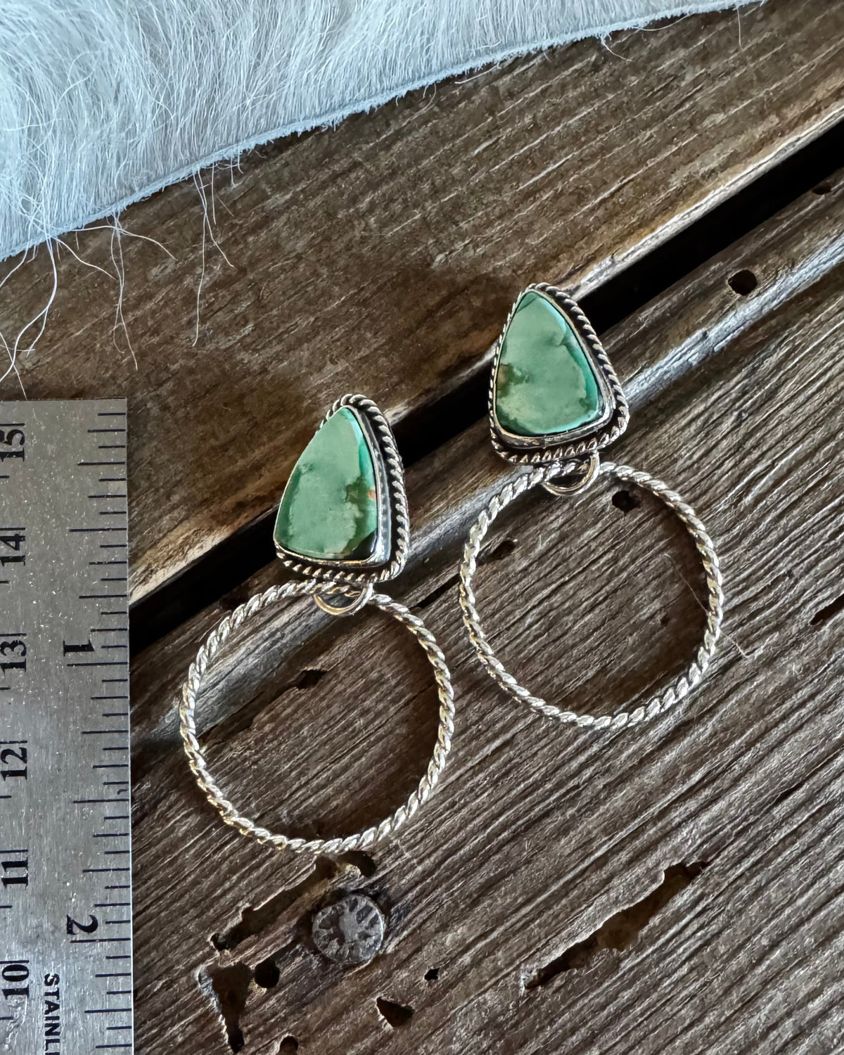 A pair of silver-toned dangle earrings with triangular light green gemstone tops and textured circular wire hoops, displayed on a rustic wooden surface next to a metal ruler for scale.