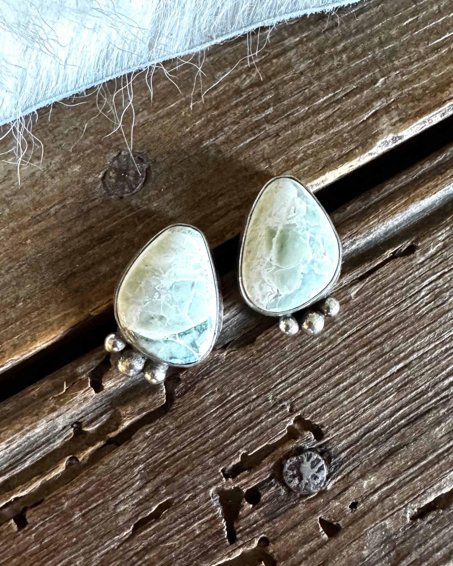 Tear-shaped light green gemstone stud earrings with silver settings and small dangling silver beads, displayed on a rustic wooden surface with a white faux fur accent.