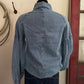 A light blue denim shirt-jacket with long puff sleeves, shown from the back on a mannequin torso with dark blue jeans. The jacket has a central seam down the back, gathered shoulder seams, and patch pockets.