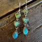 A pair of handmade dangle earrings with triangular green and turquoise stones in silver settings, displayed on a rustic dark wood background.