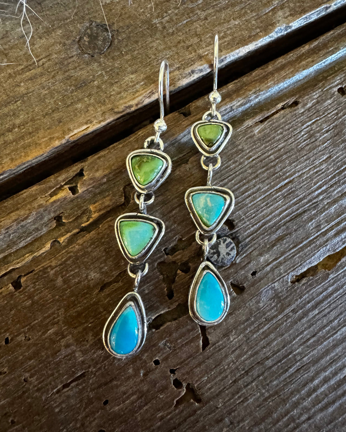 A pair of handmade dangle earrings with triangular green and turquoise stones in silver settings, displayed on a rustic dark wood background.