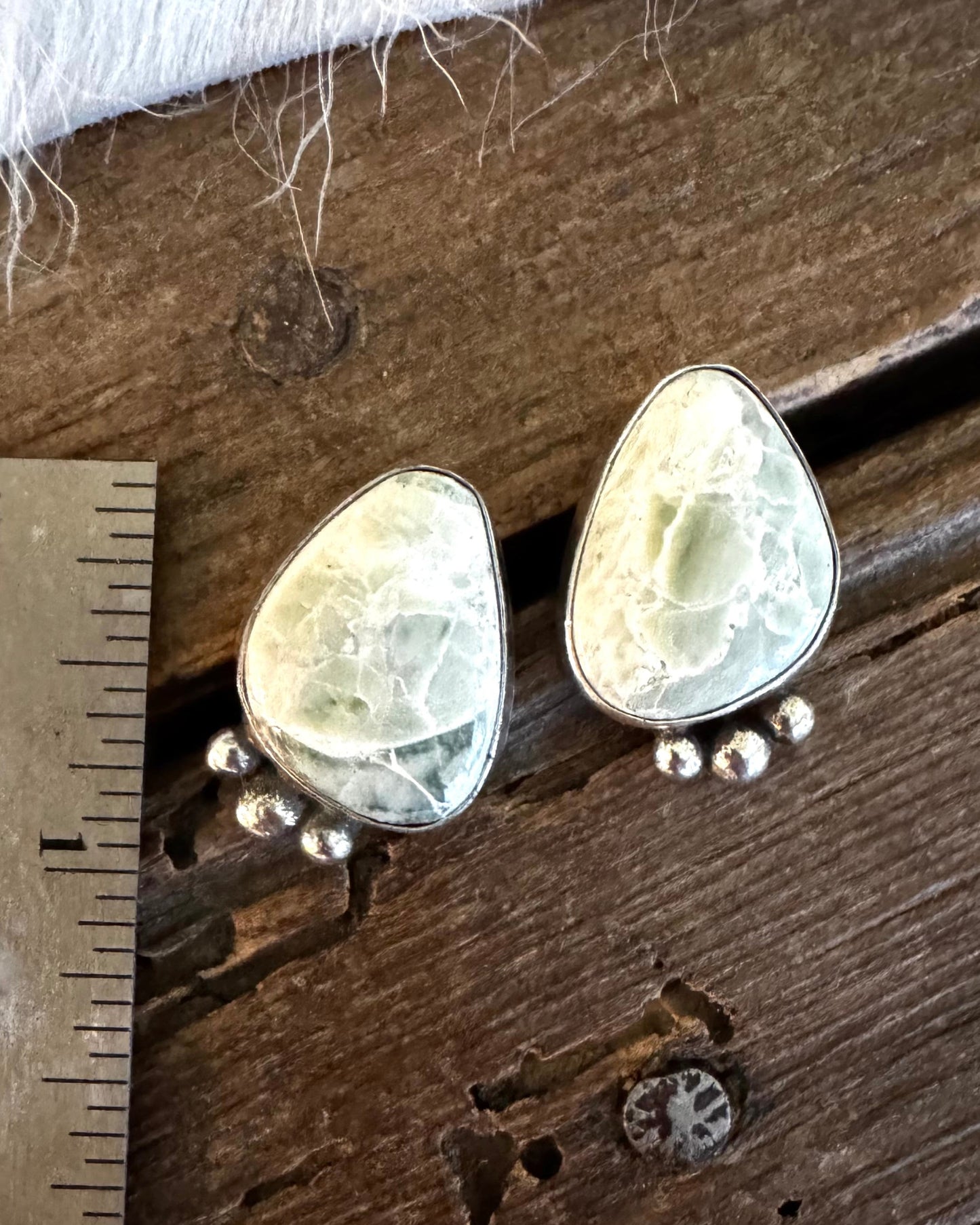 A pair of handmade stud earrings with irregular light green stone pieces in silver settings, shown on a rustic wooden surface next to a metal ruler for scale.