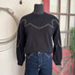 Black sweater with white stitching on a mannequin against a decorative wall.