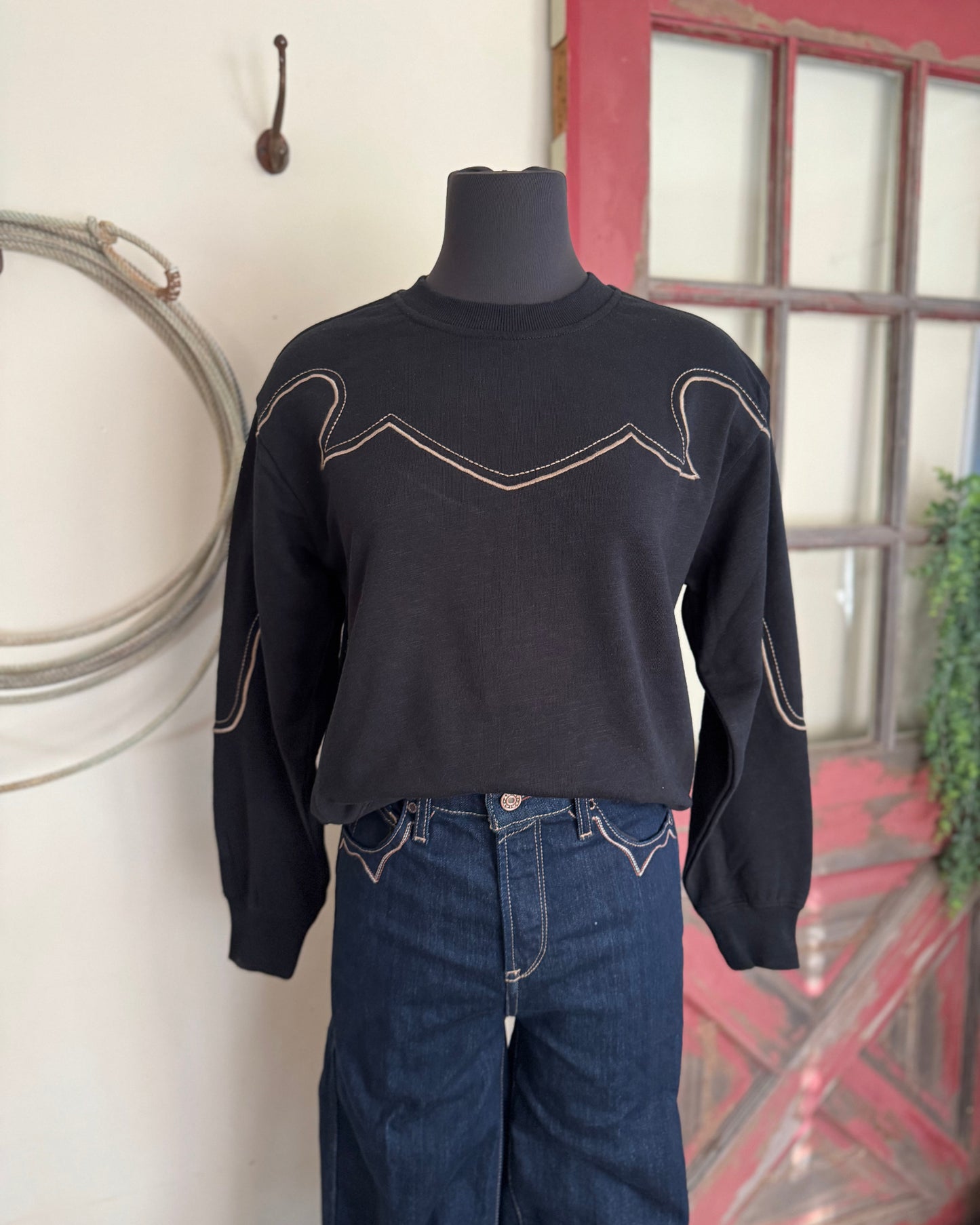 Black sweater with white stitching on a mannequin against a decorative wall.