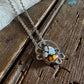 A silver-toned necklace with a circular pendant featuring white, black, and orange marbled stones, displayed on a rustic wooden surface with a white faux fur accent.