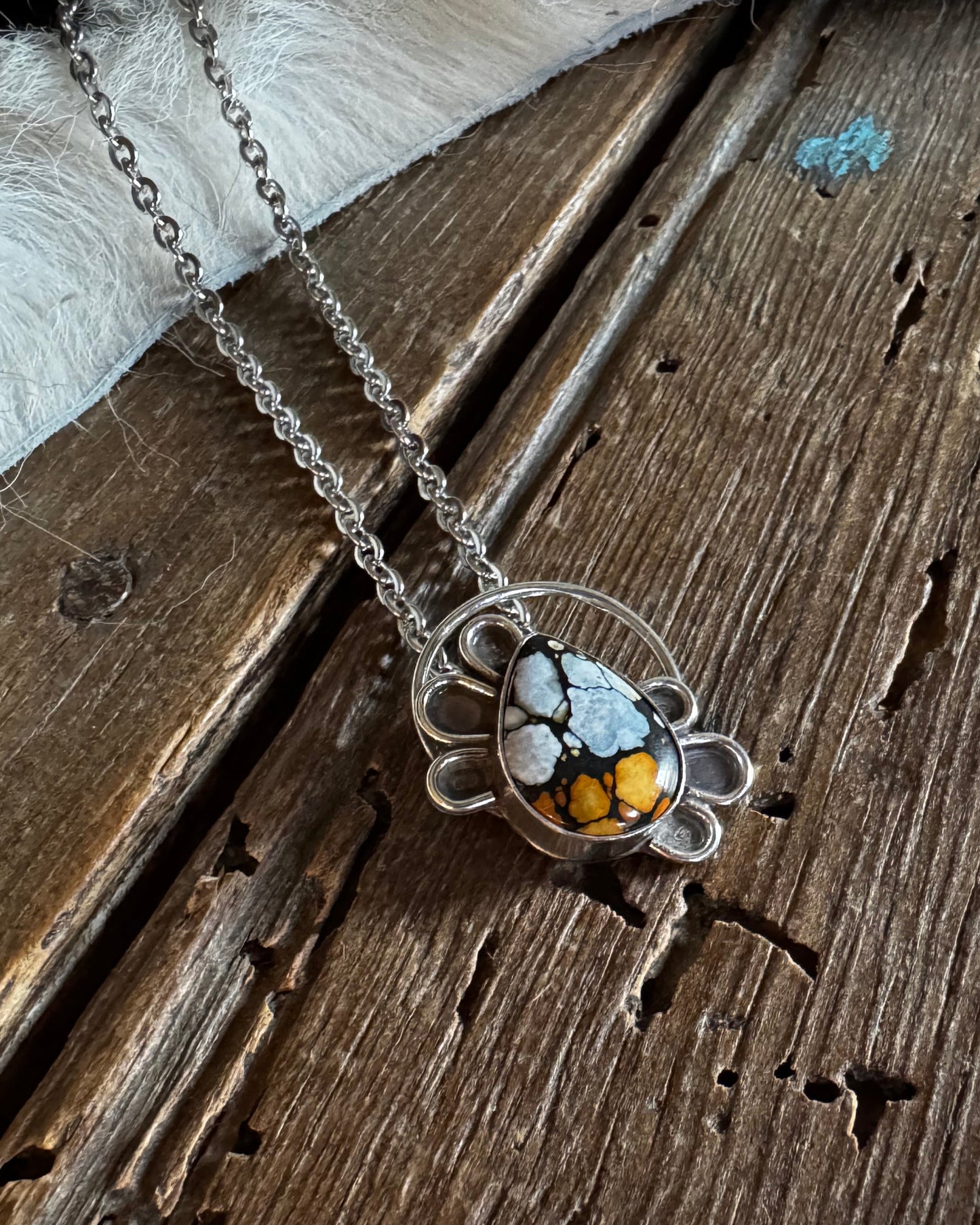 A silver-toned necklace with a circular pendant featuring white, black, and orange marbled stones, displayed on a rustic wooden surface with a white faux fur accent.