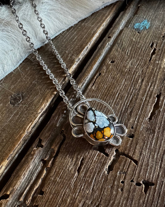 A silver-toned necklace with a circular pendant featuring white, black, and orange marbled stones, displayed on a rustic wooden surface with a white faux fur accent.
