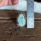 A hand holding a metal ruler next to a silver-toned ring with an oval turquoise-blue stone on a rustic wooden surface.
