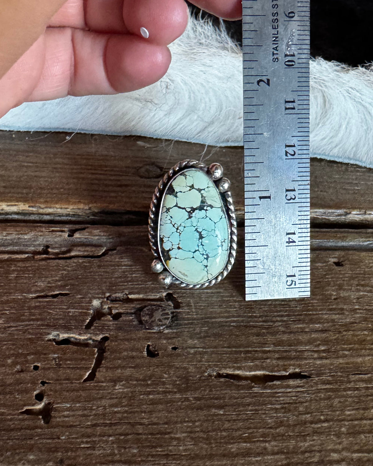 A hand holding a metal ruler next to a silver-toned ring with an oval turquoise-blue stone on a rustic wooden surface.