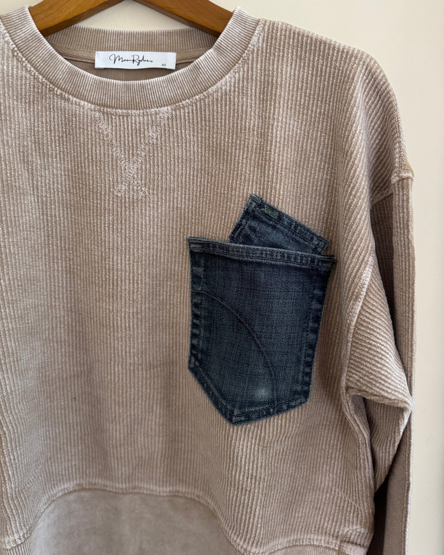 Beige ribbed sweater with a blue pocket on a hanger against a white background
