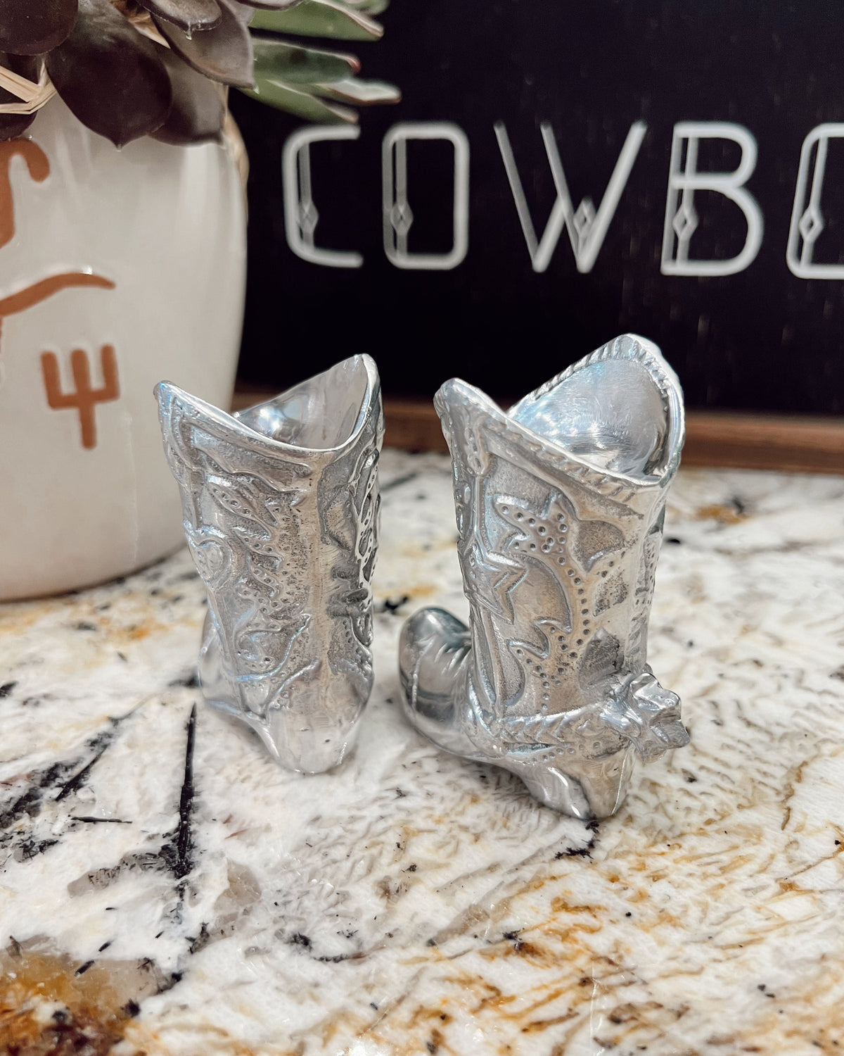 Silver cowboy boot salt and pepper shakers on a marble surface with a 'Cowboy' sign in the background.