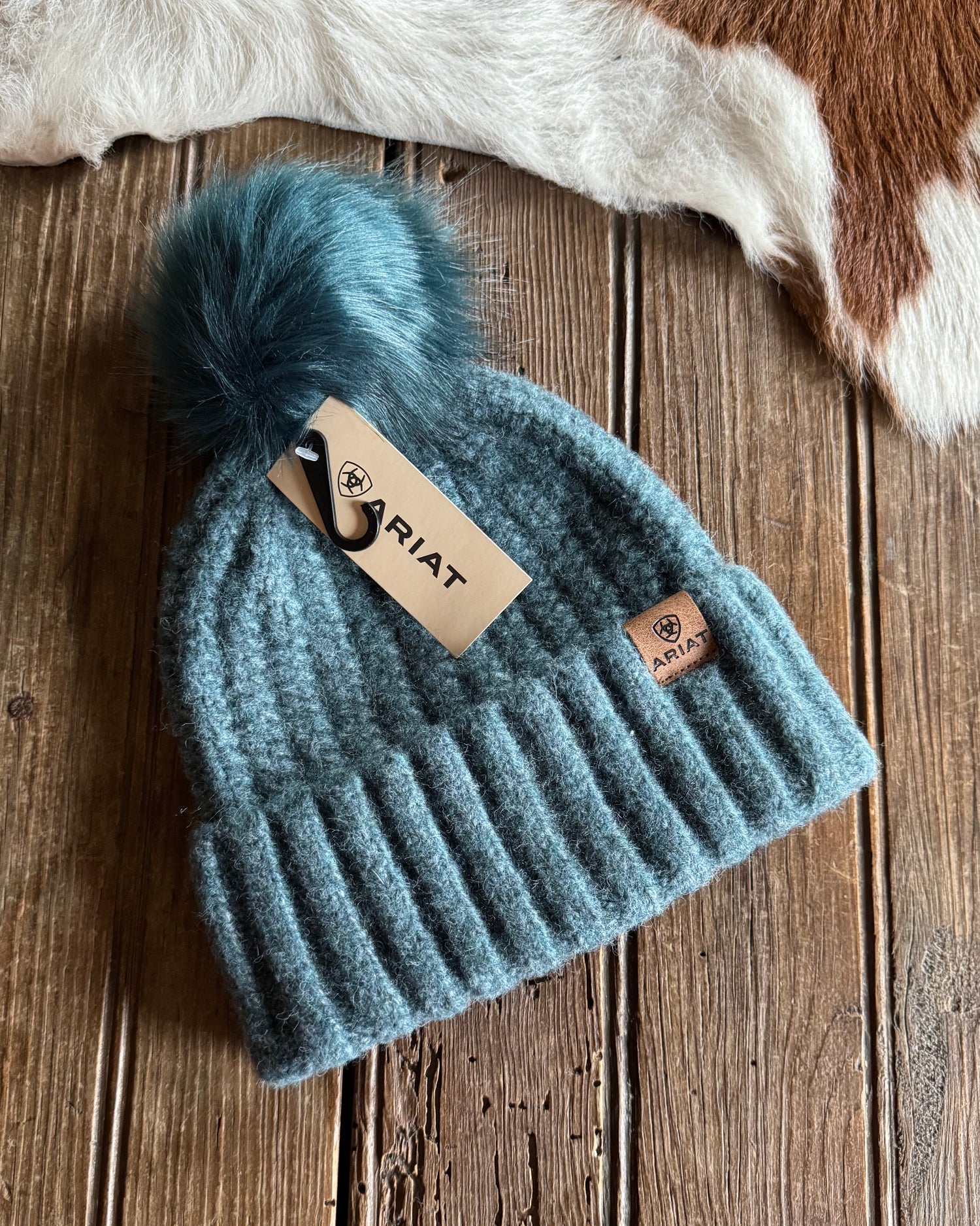Blue knit beanie with pom-pom on a wooden surface, Ariat brand visible