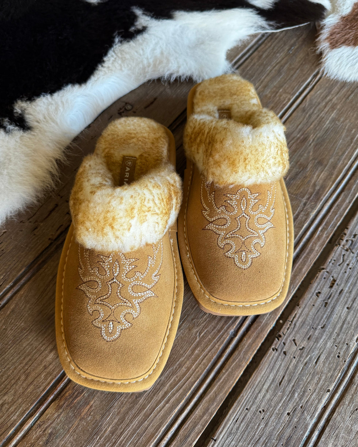 A pair of tan suede slip-on slippers with white embroidered floral patterns and fluffy cream faux-fur trim, displayed on a rustic wooden plank floor next to a black and white cowhide rug.