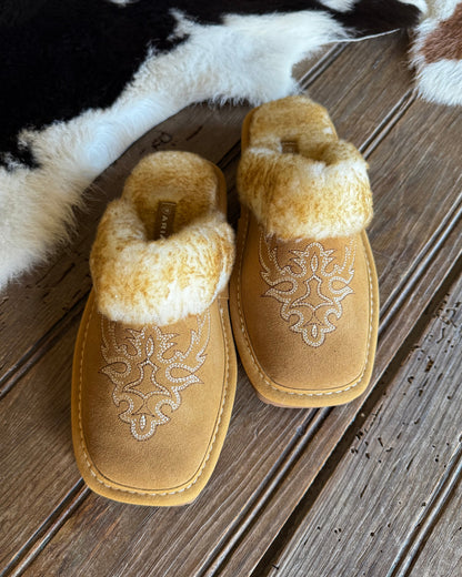 A pair of tan suede slip-on slippers with white embroidered floral patterns and fluffy cream faux-fur trim, displayed on a rustic wooden plank floor next to a black and white cowhide rug.