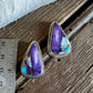 A pair of triangular turquoise and purple stone earrings with silver-toned settings, displayed on a rustic wooden surface next to a metal ruler for scale.