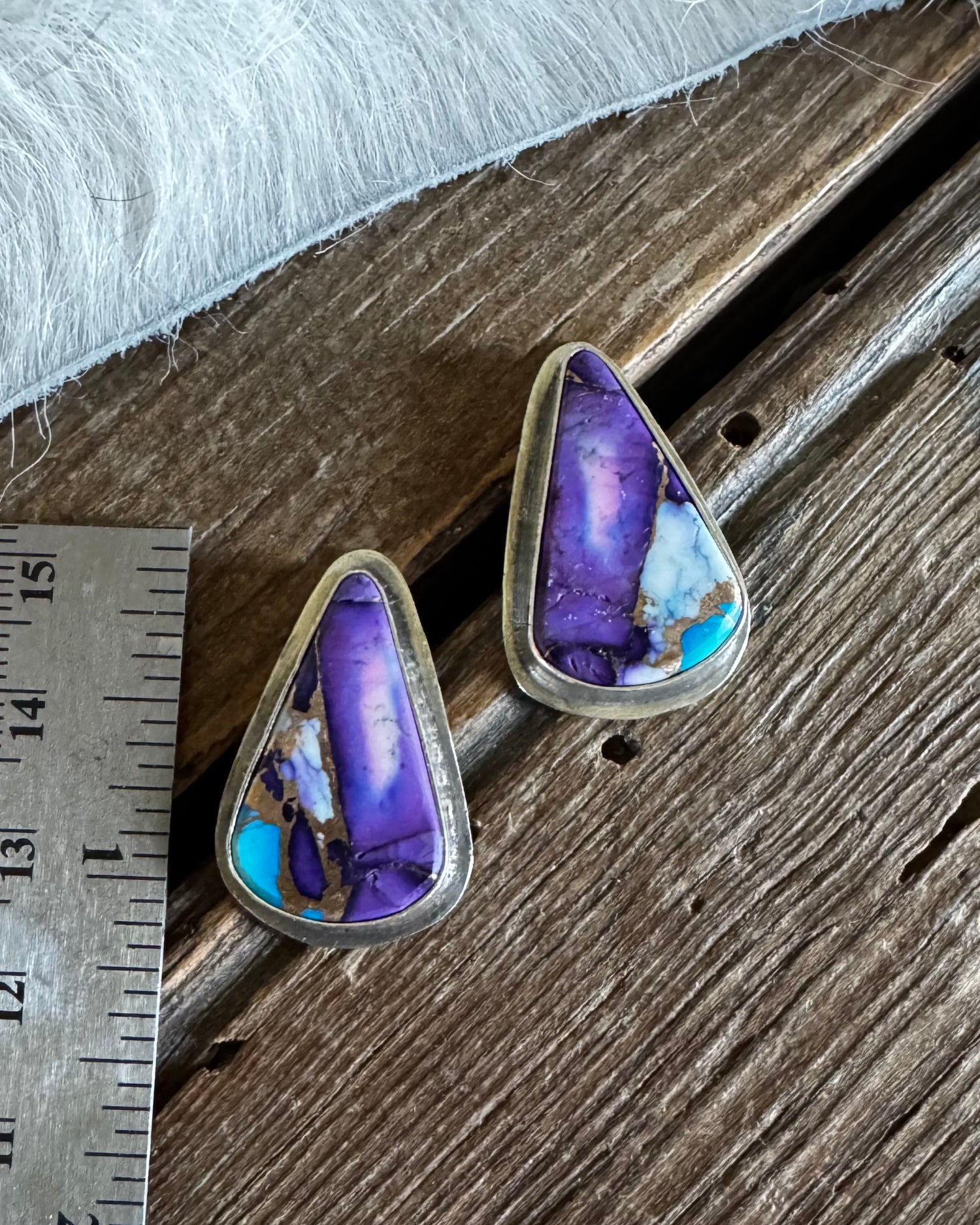 A pair of triangular turquoise and purple stone earrings with silver-toned settings, displayed on a rustic wooden surface next to a metal ruler for scale.