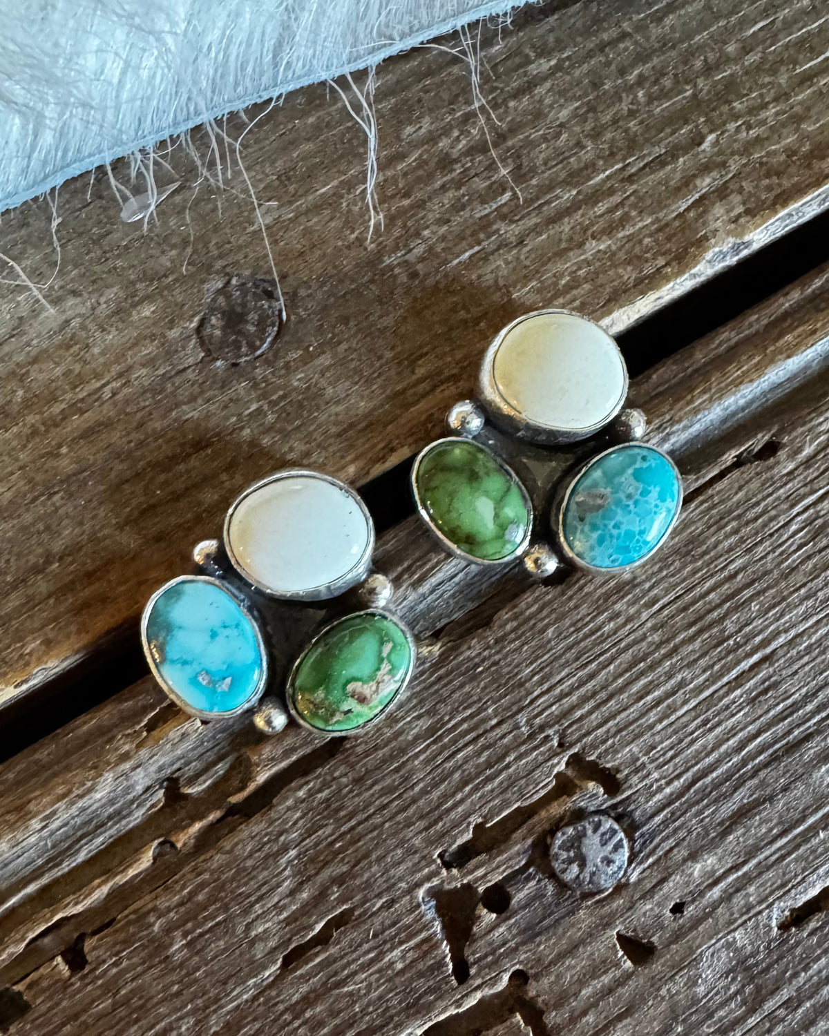 A pair of silver stud earrings with oval turquoise and green cabochon stones, displayed on a rustic wooden surface.